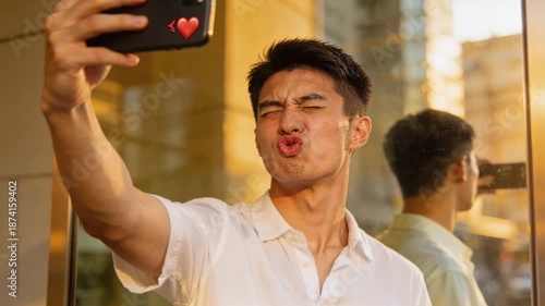 Young man smartphone reflection window city golden hour valentine smiling relaxed taking selfie near reflective glass wall