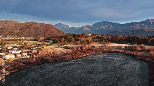 Aerial drone shot with a gentle circular movement around a frozen lake in North of Italy, creating parallax between winter trees, countryside and distant mountains at sunrise.
