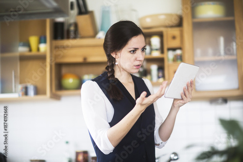 Side view of a serious Woman standing in her kitchen video conferencing on a digital tablet