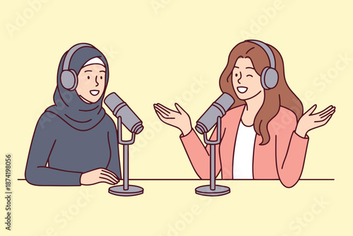 Two young women including girl in hijab sitting in studio and recording happy conversation. Inclusive female creators communicating and sharing ideas for digital audio podcast.