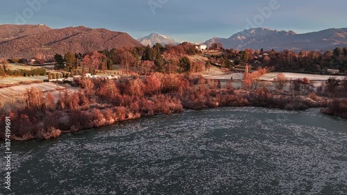 Aerial drone shot moving sideways from left to right over a frozen lake in North Italy, revealing winter countryside, bare trees and distant mountains at sunrise.