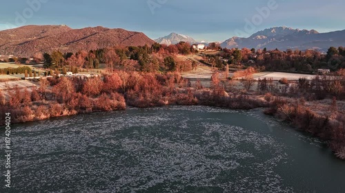 Aerial drone shot with a gentle circular movement around a frozen lake, creating parallax between winter trees, countryside and distant mountains at sunrise.