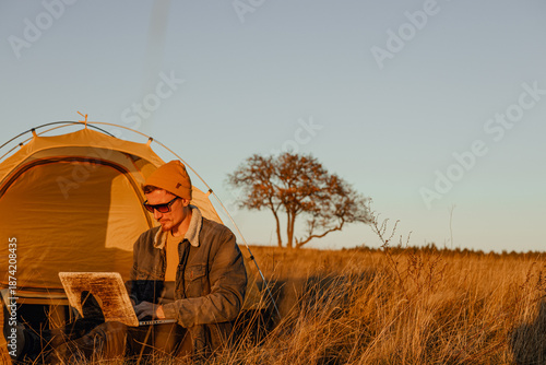 Male digital nomad using computer and working online while sitting near camping tent at campsite in nature  