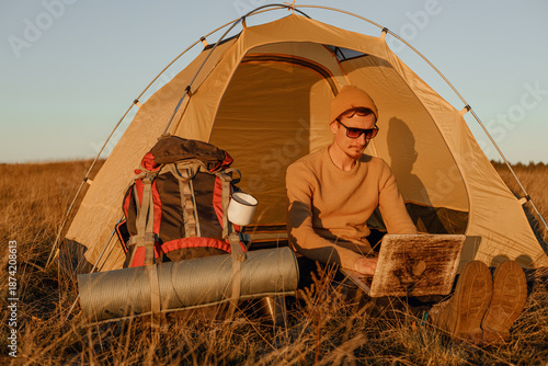 Male traveler browsing laptop during remote work while sitting near camping tent at campsite in nature at sunset  