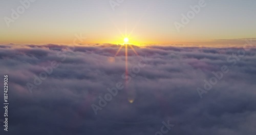 Aerial flight above glowing clouds at sunset with golden sunlight breaking through. Cinematic skyscape with soft fog, warm colors and dramatic atmosphere high above the horizon.