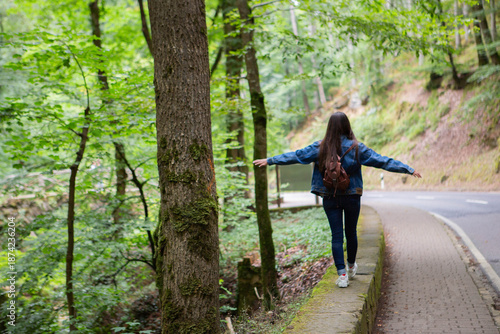 Woman back silhouette in denim walking on asphalt road through green forest balancing