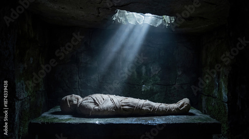 Body of Jesus wrapped in linen shroud lying in dark stone tomb. Ray of divine light shining from above on burial place. Biblical concept of Holy Saturday and impending Resurrection.