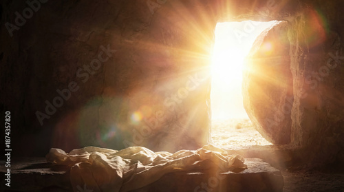 Empty tomb of Jesus with rolled stone and burial linens on slab. Bright divine light streaming into dark cave. Biblical concept of Resurrection miracle and Easter Sunday victory.