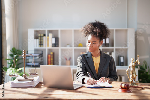 Young Black woman attorney working on documents, surrounded by legal instruments like scales of justice, a gavel, and a laptop, symbolizing professionalism, law, and business