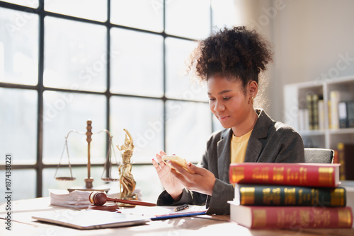 Young female law student reviewing legal documents at an outdoor desk with law books, scales of justice and gavel nearby, using a smartphone while focused on study and research