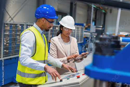 Female manager guides male worker on production process in busy factory. Focus on collaboration and workplace communication.