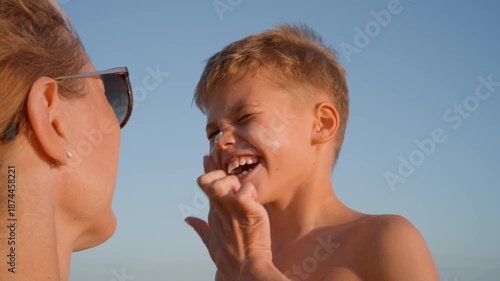 Close up of a mother applying sunblock cream on her smiling son's face during a summer holiday on a sunny beach. Health and skin protection concept.