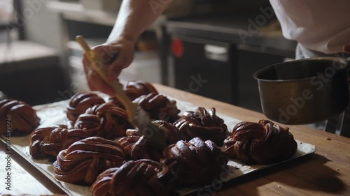 Baker glazing freshly baked cinnamon buns with sweet syrup. Golden crust shines as the syrup slowly absorbs into the warm pastry, artisan quality handcrafted bakery. Hygge atmosphere, premium bakery.