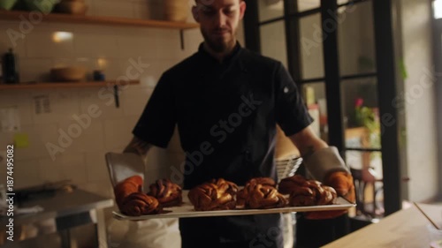 A baker looks at the camera and smiles, holds a tray of freshly baked buns in a cozy artisan bakery.Warm lighting, professional work in the kitchen, fresh bread and buns ready for sale.Factory worker.