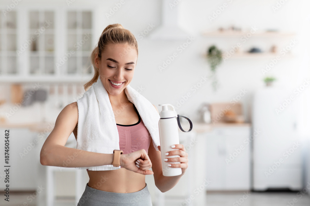 Fototapeta premium Smiling caucasian pretty young blonde woman checking heartbeat on fitness tracker and drinking water after workout at home, empty space. Training with gadget, body and health care, aqua balance indoor