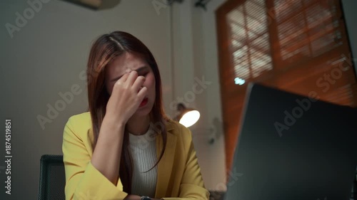 Young Asian business professional feeling tired and frustrated, massaging her temples due to eye strain and stress from overworking at night
