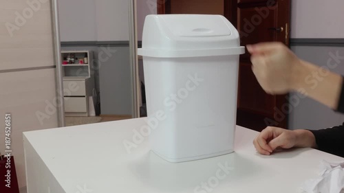 Adult woman hands throwing crumpled paper into white plastic trash bin indoors, symbolizing waste disposal, rejection, cleanup and environmental concept.