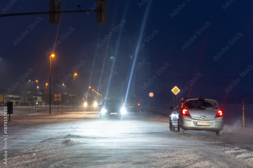 Fototapeta premium Extreme winter weather night with heavy snowstorm strong wind icy road surface truck cars driving blizzard bright headlights cutting low visibility. Dangerous urban traffic conditions Germany