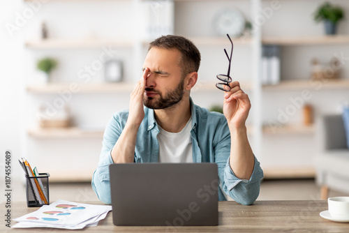 Fotografie Caucasian male freelancer feeling tired, rubbing irritated eyes, sitting at desk with laptop, exhausted from online work