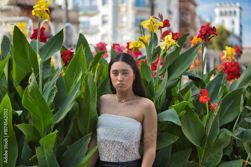 Young woman, attractive, brunette, blue eyes, Hispanic, wearing a white top, posing and looking at the camera surrounded by plants with beautiful colorful flowers. Concept: model, beauty, fashion.