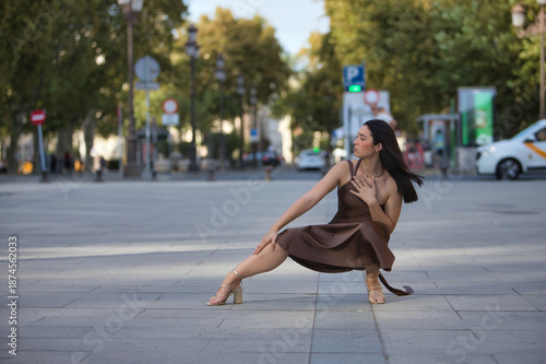 Young woman, beautiful, brunette, blue eyes, Hispanic, wearing an elegant brown chiffon dress and gold heels, posing crouched on a city sidewalk while the wind blows her hair. Concept: model, beauty.