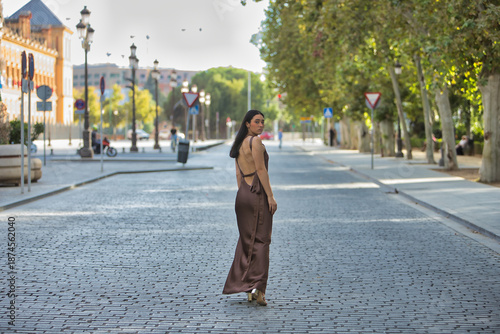 Young woman, beautiful, brunette, blue eyes, Hispanic, wearing an elegant brown chiffon dress and gold stiletto heels, walking down a cobbled street in the city. Concept: model, beauty, fashion.