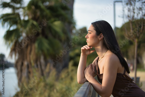 Young woman, beautiful, brunette, blue eyes, Hispanic, wearing an elegant brown chiffon dress, with her hand on her chin, gazing into the distance, leaning on a metal railing. Concept: model, beauty.