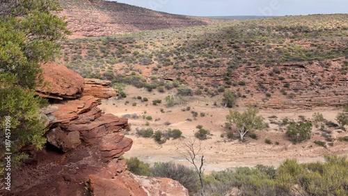 A wide view shows rocky formations, sparse bushes, and a dry riverbed in a dry area in Australian outback