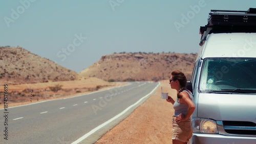 Young adult woman leans against a van on a long, empty road surrounded by brown hills while holding a cup