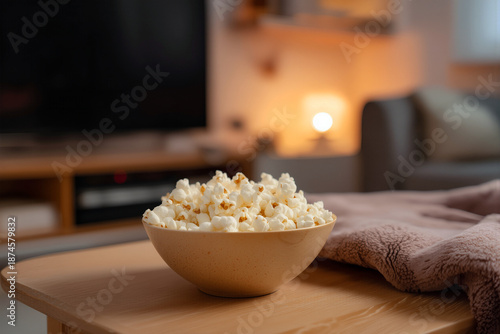 Cozy home movie night with bowl of popcorn and warm blanket on wooden table. National Popcorn Day celebration in living room with soft lighting and TV on blurred background.