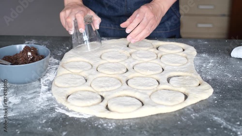 Person prepares cookie dough on a kitchen counter. Round pieces of dough are being cut using a cutter. Flour is spread around for easy handling. Ingredients are nearby. 