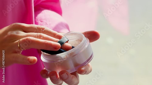 Closeup of a beautician holding a jar of loose setting powder with a puff, preparing to apply makeup in a salon. Bright daylight, clean tools, professional beauty service