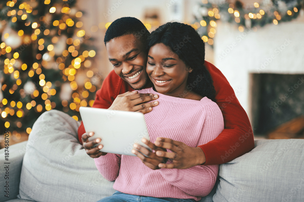 Fototapeta premium A couple sits together on a gray sofa, smiling and looking at a tablet. The room is decorated for the holiday season with lights and a Christmas tree in the background.