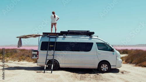 Young adult woman looks out over pink lake from the roof of a parked van on a sunny day, Western Australia