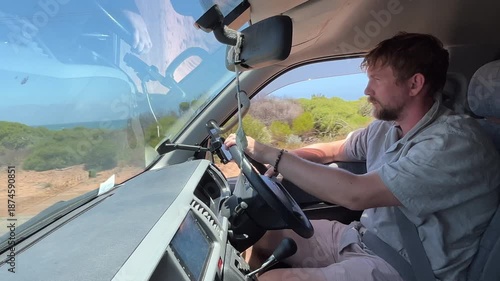 Yound adult man drives a vehicle along a coastal road, gazing at the ocean and enjoying the view of the water and land.