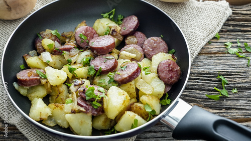 Sausage and potatoes are cooking together in a frying pan. The dish looks fresh with green herbs on top. The setting includes a wooden table and a textured cloth.