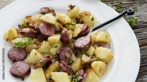Dish of cooked potatoes and sausages served on a white plate with green herbs on wooden table