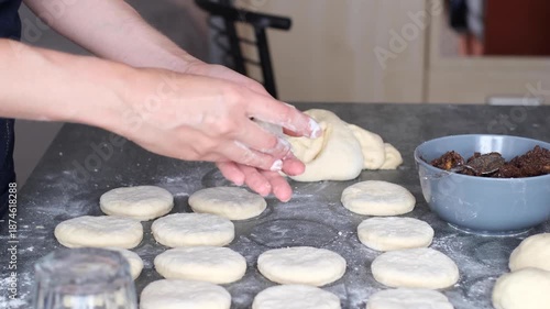 Hands preparing poppy seed buns on a kitchen table. Small pieces of dough put poppy seed filling are shaped into round forms. A bowl of filling sits nearby on the table. side view