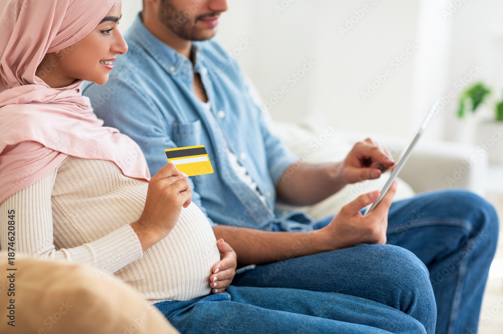 Obraz premium Online shopping for pregnant couple. Muslim man and woman using digital tablet and credit card, buying things for their future baby, sitting together on sofa