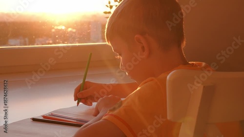 Focused young student. Young boy concentrates attentively on his schoolwork amid sunlight and city view. Young child carefully drawing colorful pencils while sunlight illuminates room and city skyline