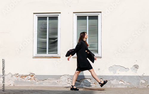 Εκτύπωση καμβά Young woman in black dress and sandals walks confidently along a textured wall,