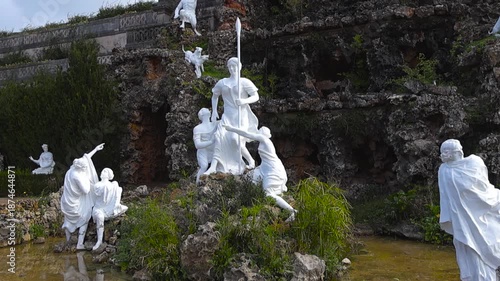Detailed view of white classical statues in a rocky grotto fountain at Quinta Real de Caxias, surrounded by greenery and shallow water in a historic garden setting.