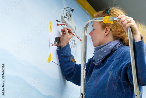 Female artist painting a mural on an exterior wall while standing on a ladder, focused creative work process with brush and reference sketch, representing creativity and professional wall art