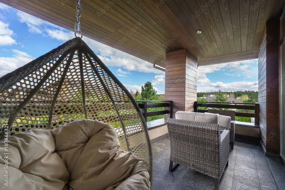 Obraz premium A balcony features a comfortable hanging chair and a seating arrangement. Surrounding greenery and distant structures are visible under a bright sky with clouds.