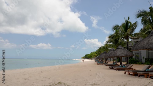 Luxury tropical resort beach with soft white sand, green palm trees, and straw umbrellas under a bright blue sky with white clouds. Relaxing atmosphere at a premium beach club 