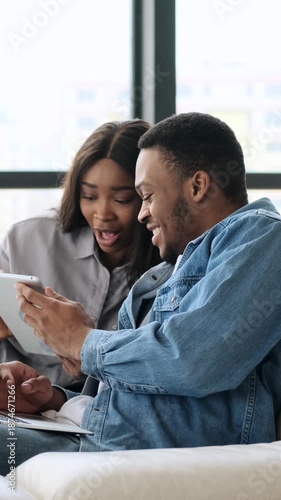 Vertical video of a happy young African American couple using a tablet and laptop together while sitting on a couch at home.