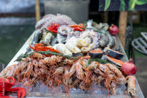 Assortment of raw seafood on ice at outdoor fish market, including shrimp, squid, octopus, mussels, salmon and crab. Fresh catch display with herbs and pomegranate.