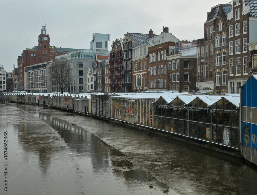 Fototapeta premium Amsterdam’s Bloemenmarkt floating flower market in a rare winter scene. The canal surface is icy, but main waterways rarely freeze solid enough for skating
