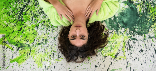 Young woman lies on a color splashed surface with vivid green and black paint. She looks up with calm eyes and soft expression, wrapped in a fluffy bathrobe. Vibrant and expressive mood