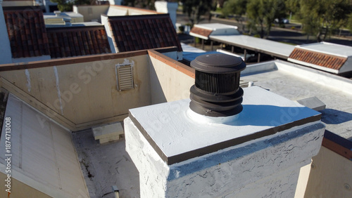 Flat roof chimney with dark metal rain cap mounted on sealed white masonry structure, surrounded by stucco walls and clay tile rooftops within a residential housing complex